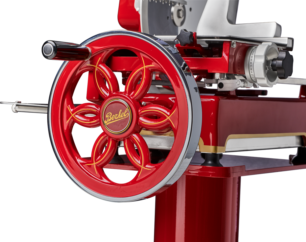Close-up of a vintage red Berkel Fly Wheel Slicer, highlighting the ornate handwheel with floral spokes and yellow lettering, chrome edge, and part of the slicing mechanism with a chromium-plated carbon steel knife. Polished metal details complete its glossy red look.