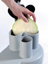 A hand places a halved green cabbage into the gray cylindrical chute of a Robot Coupe R301 Dice combination food processor, preparing it for slicing or shredding. The plain white background highlights the focus on the cabbage and device.