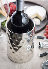 A dark wine bottle in a stainless steel, double wall insulation wine cooler sits on a table beside a corkscrew, grapes, and brie cheese. A wine glass and blurred background items suggest an inviting wine and cheese setting.