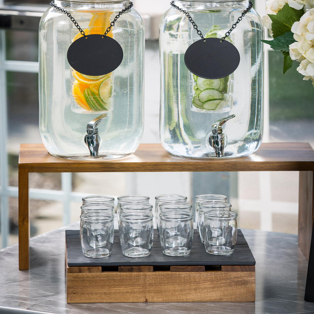 Two large glass drink dispensers with blank black labels sit on an acacia wood nesting set. One holds water with citrus slices; the other has cucumber water. Below, nine empty glass cups are arranged on a wooden tray.