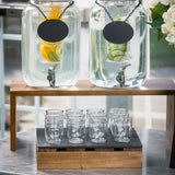 Two large glass drink dispensers with blank black labels sit on an acacia wood nesting set. One holds water with citrus slices; the other has cucumber water. Below, nine empty glass cups are arranged on a wooden tray.