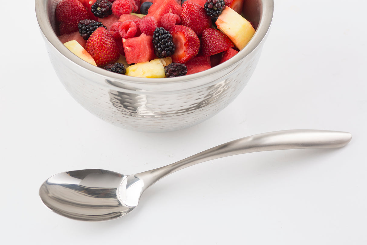 A silver bowl from the Dalton II Collection filled with assorted fresh fruit pieces, including strawberries, pineapple, blackberries, and watermelon, sits on a white surface beside a shiny stainless steel serving spoon.