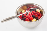 A stainless steel bowl from the Dalton II Collection is filled with assorted fresh fruit—strawberries, raspberries, blackberries, pineapple, and watermelon. A metal serving spoon rests elegantly inside on a white surface.