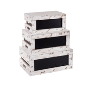 Three weathered, white wooden crates with black chalkboard panels are stacked as a Rustic Risers Crate Set. Each distressed wood crate has rectangular cut-out handles and a vintage look, perfect for adding rustic charm. The background is plain white.