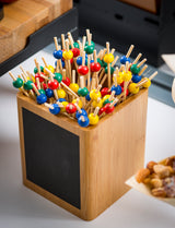 A bamboo storage container with a blank blackboard label holds skewers topped with colorful beads in red, yellow, blue, and green. Part of a wooden tray and a dish of assorted nuts are visible nearby in the background.