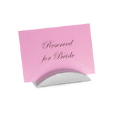 A pink card with elegant cursive writing reading Reserved for Bride is displayed upright in a sleek TableCraft stainless steel card holder against a plain white background.