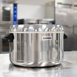 A large, stainless steel Zojirushi thermal rice cooker with black handles sits on a shiny metal countertop in a commercial kitchen, with blurred ovens and appliances visible in the background.