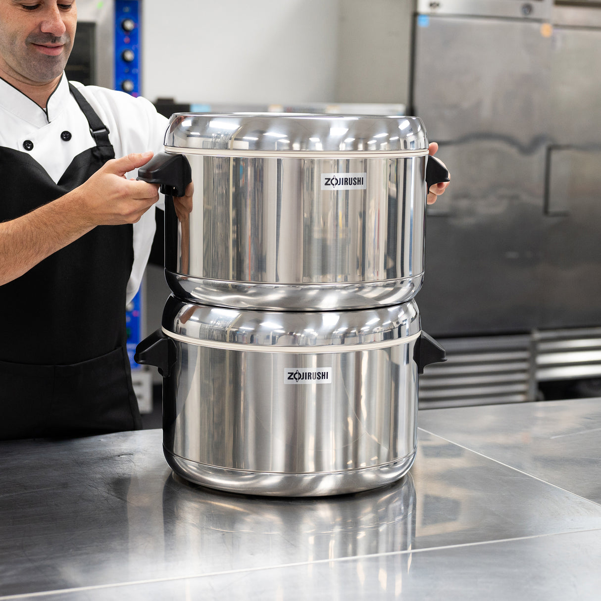 A person in a chef uniform stands in a kitchen, holding the handles of two large, stacked Zojirushi thermal rice cookers—ideal for keeping sushi rice fresh—on a reflective metal countertop. Industrial appliances and cabinets fill the background.