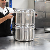 A person in a chef uniform stands in a kitchen, holding the handles of two large, stacked Zojirushi thermal rice cookers—ideal for keeping sushi rice fresh—on a reflective metal countertop. Industrial appliances and cabinets fill the background.
