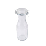 A clear glass carafe with a wide neck and round body, topped with a hinged metal clasp and airtight glass lid, stands upright on a white background. This 34 oz carafe is perfect for serving water or juices in style.