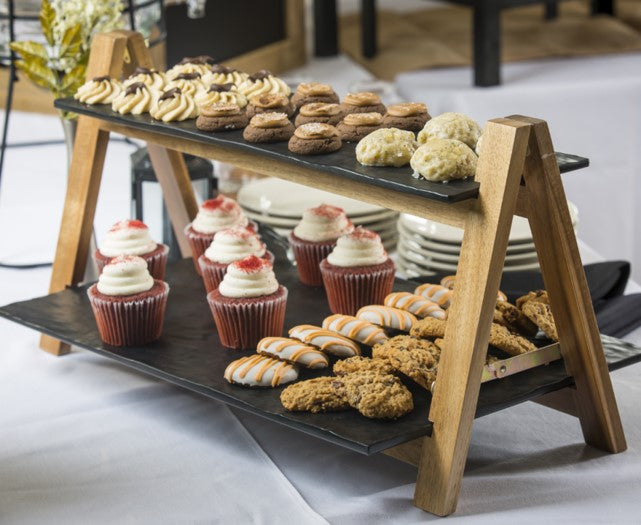 An acacia wood riser from the TableCraft Products Frostone Collection holds assorted pastries: cookies and swirled mini tarts on top, cupcakes with white frosting, striped cookies, and oatmeal cookies below. Plates are stacked on a white tablecloth nearby.