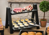 A black two-tiered stand from the Frostone Collection holds rolled cured meats with cheese on top and deviled eggs below. A pair of tongs sits beside the eggs, while a small potted plant adds charm against a stone wall.