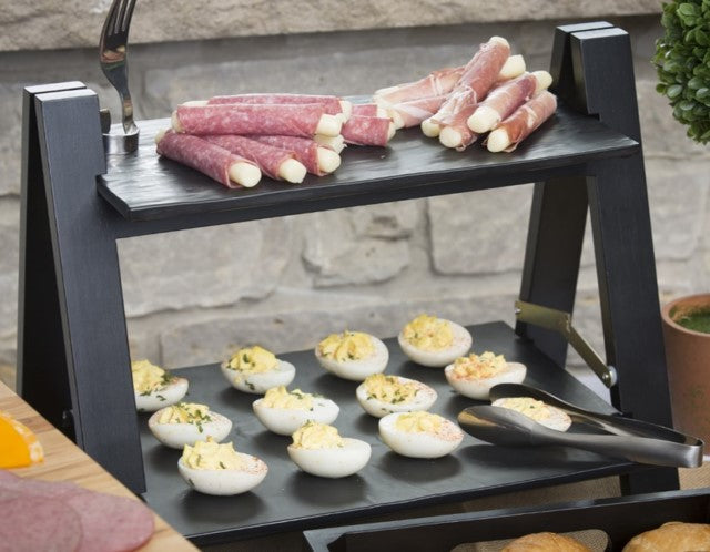 Two-tiered black serving tray displayed on a TableCraft Products Frostone Riser Frame, with rolled deli meats and cheese sticks on top and deviled eggs with herbs below. Metal tongs rest on the lower shelf; a stone wall forms the backdrop.