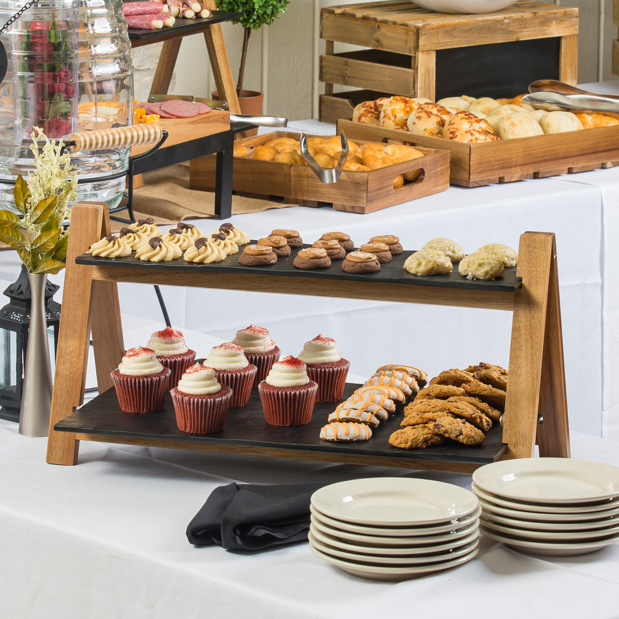 A buffet table displays assorted pastries and desserts, including cupcakes, cookies, and biscuits on a 2-tier wood riser. Plates are stacked in the foreground, with more baked goods on tables draped in white tablecloths.