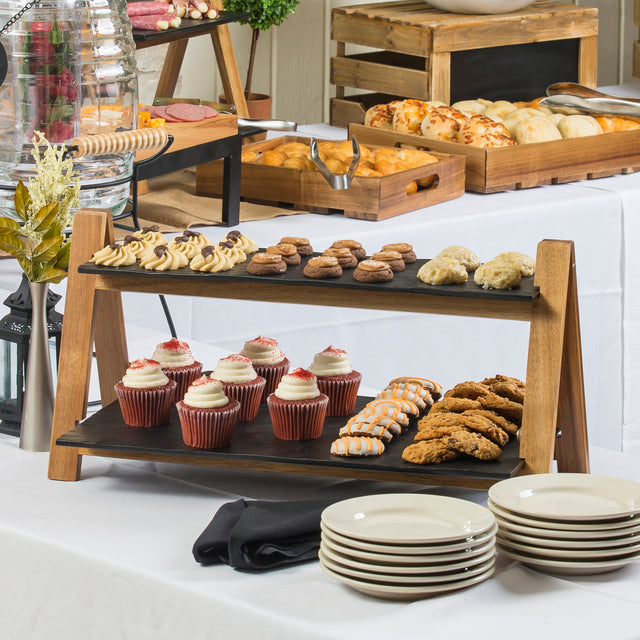 A buffet table displays assorted pastries and desserts, including cupcakes, cookies, and biscuits on a 2-tier wood riser. Plates are stacked in the foreground, with more baked goods on tables draped in white tablecloths.