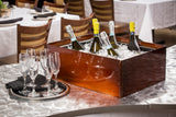 A Remington Collection Designer Tote Box filled with ice holds six bottles of sparkling wine. Next to it, empty champagne flutes rest on a silver tray atop a modern metallic table in a restaurant with neatly set tables and chairs in the background.