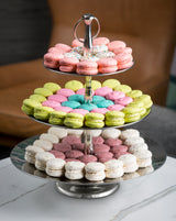 A three-tiered silver stand from the Remington Collection Serving Set displays colorful macarons in circles. Pink, green, purple, blue, and white treats are arranged on the stainless steel serving set atop a marble table.