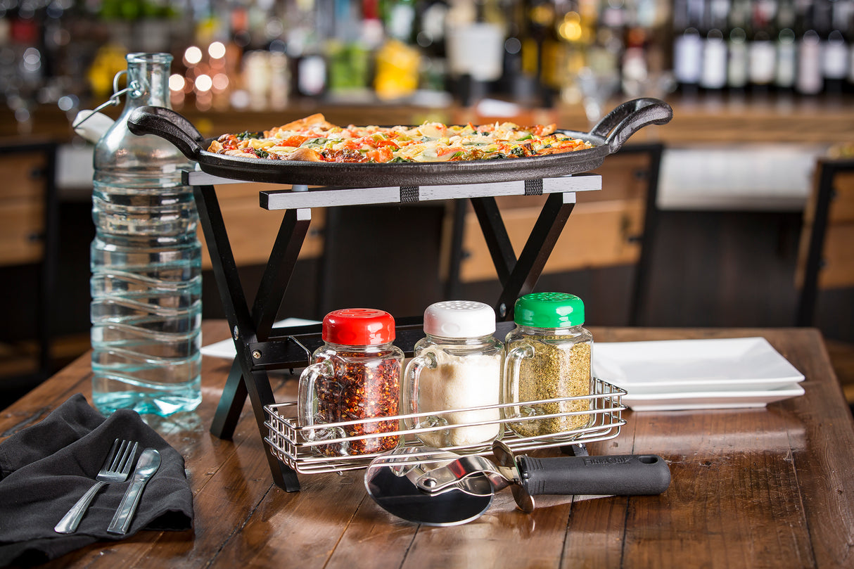 A pizza sits on a black, stainless steel rack above a wooden table set with bottled water, napkin, cutlery, three shakers, a pizza cutter, and stacked white plates in a restaurant with a blurred bar in the background.