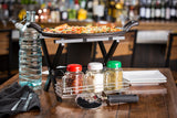 A pizza sits on a black, stainless steel rack above a wooden table set with bottled water, napkin, cutlery, three shakers, a pizza cutter, and stacked white plates in a restaurant with a blurred bar in the background.