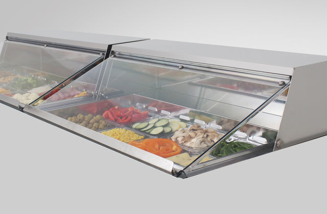 A stainless steel salad sandwich unit with a clear polycarbonate lid displays fresh ingredients—sliced bell peppers, mushrooms, cucumbers, olives, corn, and greens—neatly arranged in metal trays and kept cool with built-in refrigeration.