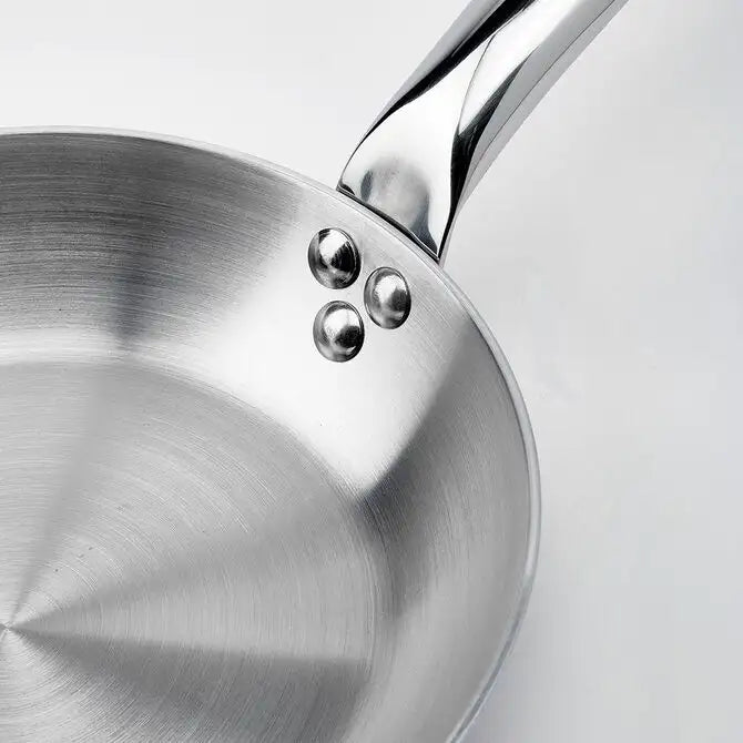 A close-up of a De Buyer Alchimy Saute Pan on a white background, showing its shiny stainless steel surface, three rivets attaching the handle, and brushed metal texture inside this 9.5 inch saute pan. The handle extends out of the upper right corner.