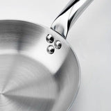 A close-up of a De Buyer Alchimy Saute Pan on a white background, showing its shiny stainless steel surface, three rivets attaching the handle, and brushed metal texture inside this 9.5 inch saute pan. The handle extends out of the upper right corner.
