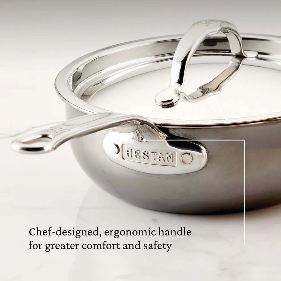 A close-up of a shiny 2.0qt Hestan Nanobond Saucier pan with a covered lid, both featuring ergonomic handles. The handle is engraved with HESTAN. The marble background highlights the chef-designed comfort and safety features.