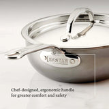 A close-up of a shiny 2.0qt Hestan Nanobond Saucier pan with a covered lid, both featuring ergonomic handles. The handle is engraved with HESTAN. The marble background highlights the chef-designed comfort and safety features.