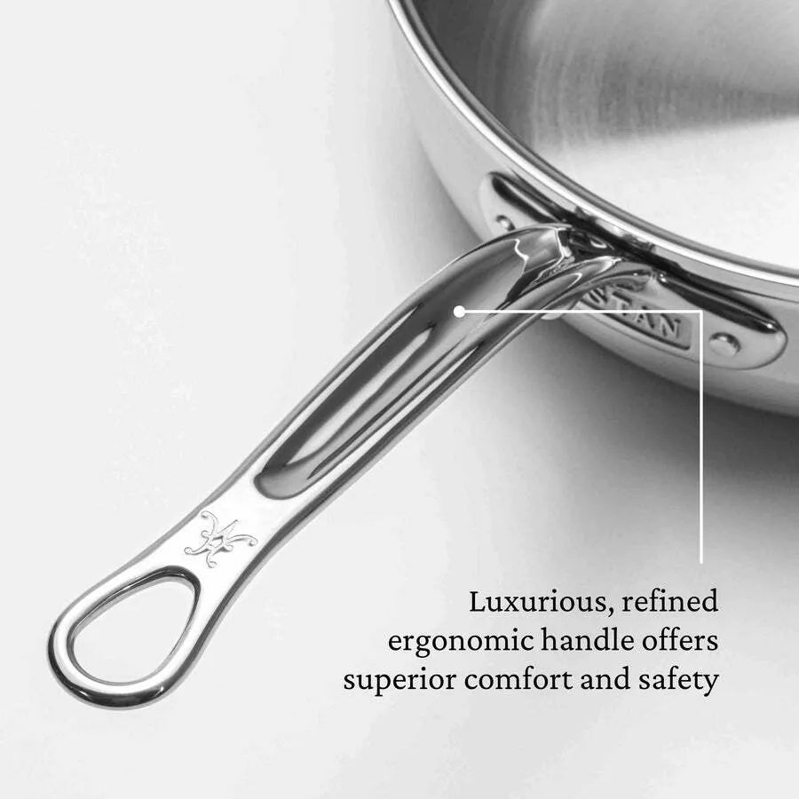 A close-up of a polished stainless steel Hestan Probond Essential Pan handle with an ergonomic, curved design. An arrow points to the handle with text: “Luxurious, refined ergonomic handle offers superior comfort and safety.” The pan’s shiny surface is visible.