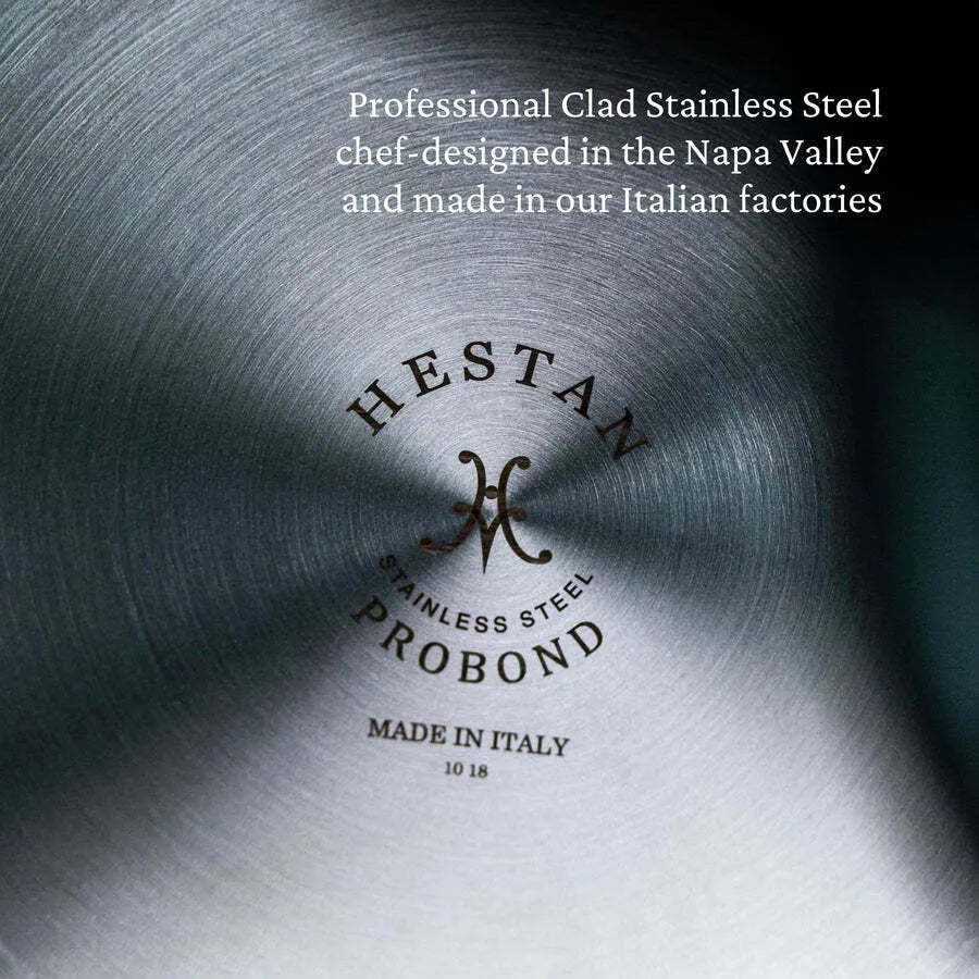 Close-up of brushed stainless steel cookware. The Hestan ProBond Essential Pan 28cm/5.0qt logo is engraved in the center, along with Stainless Steel, Made in Italy, and “10 18.” White text above reads: Chef-designed in Napa Valley, made in our Italian factories.