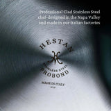 Close-up of brushed stainless steel cookware. The Hestan ProBond Essential Pan 28cm/5.0qt logo is engraved in the center, along with Stainless Steel, Made in Italy, and “10 18.” White text above reads: Chef-designed in Napa Valley, made in our Italian factories.