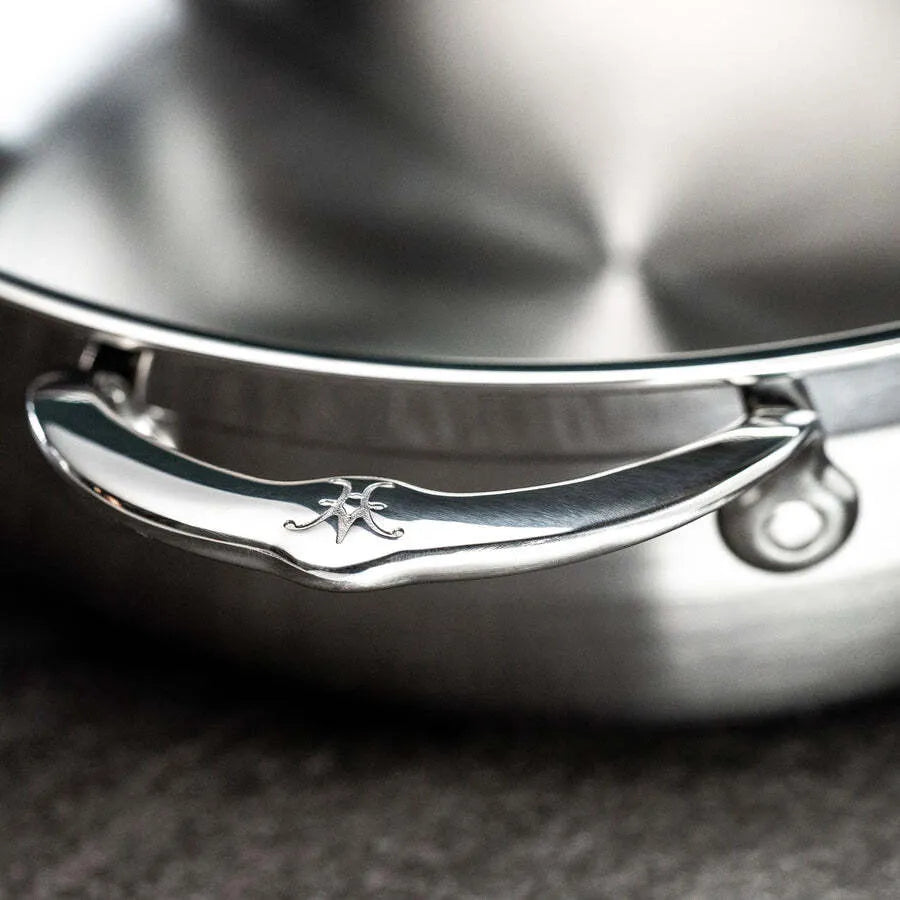 Close-up of a shiny stainless steel handle engraved with the Hestan Probond Essential pan logo, attached to a 28cm brushed metal pot lid. The background is out of focus, featuring a gray countertop surface.