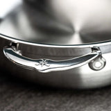 Close-up of a shiny stainless steel handle engraved with the Hestan Probond Essential pan logo, attached to a 28cm brushed metal pot lid. The background is out of focus, featuring a gray countertop surface.