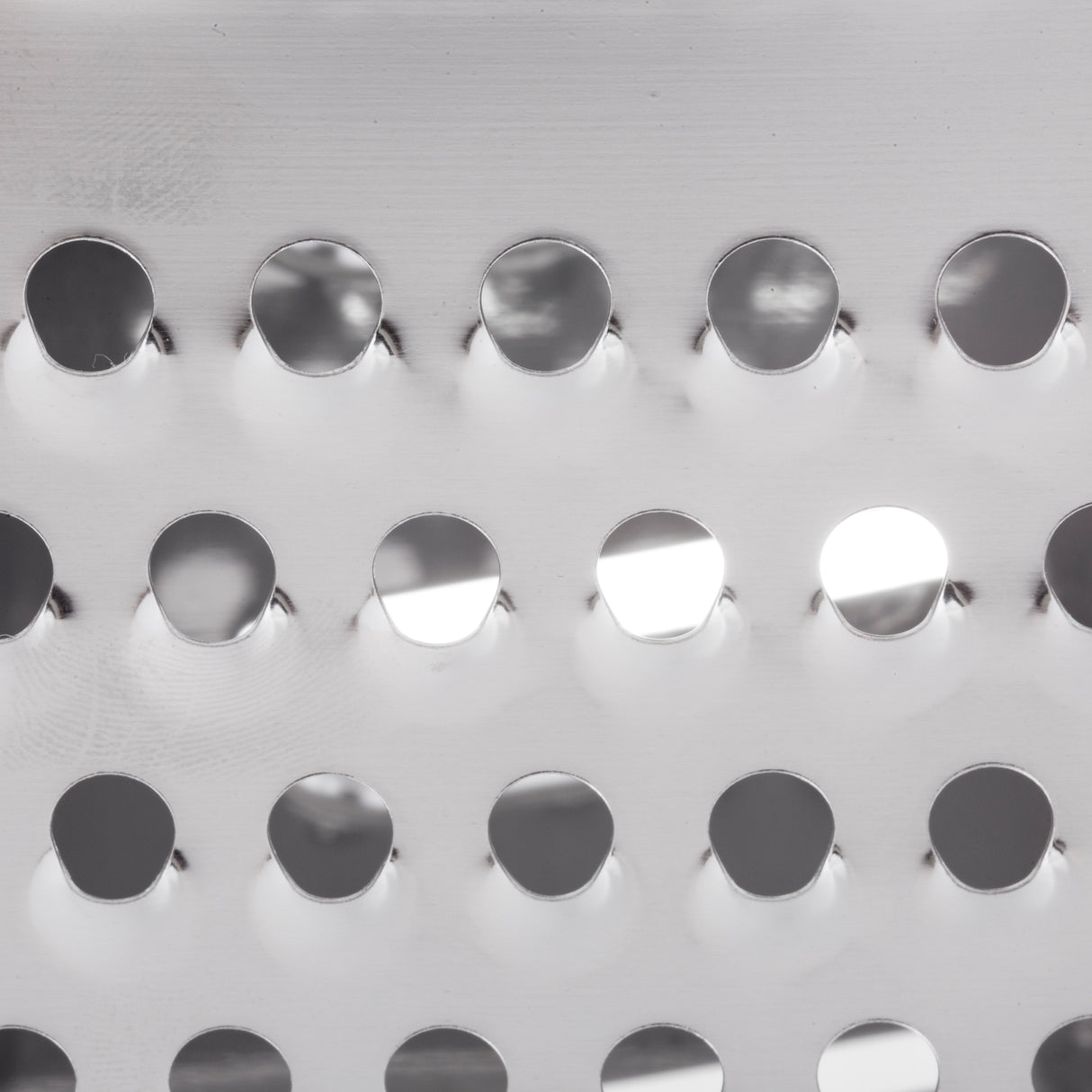 Close-up of a stainless steel surface with three horizontal rows of round holes. The shiny, reflective finish and interplay of light create highlights and shadows, giving this metal a modern, industrial look—perfect for a grater or other dishwasher safe tools.