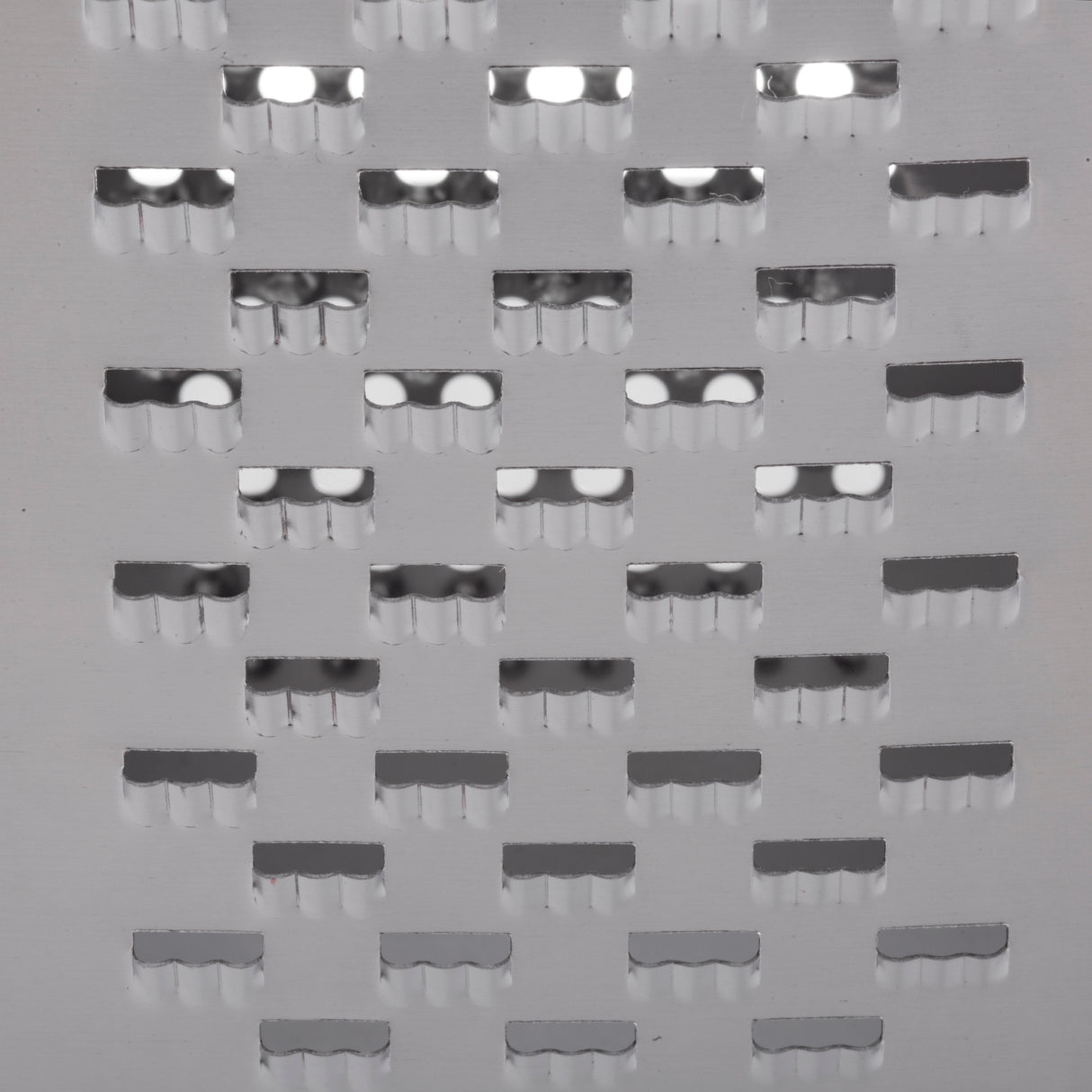 Close-up of a stainless steel grater surface with multiple rectangular holes, each edged with semi-circular notches. The shiny, silver metal reflects light, highlighting the smooth and sharp contours. Dishwasher safe for easy cleaning.