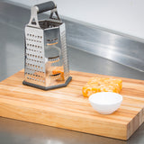 A stainless steel box grater, a block of marbled cheddar cheese, and a small empty white bowl sit on a light wooden cutting board on a kitchen counter with a stainless steel backsplash.