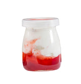 A dishwasher safe, single-serve glass jar filled with white yogurt and a layer of bright red fruit sauce at the bottom. The contents are slightly mixed where yogurt meets sauce, and the jar is sealed with a clear plastic lid on a white background.