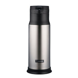 A stainless steel Zojirushi beverage server with vacuum insulation, featuring a black plastic lid and base. The upright cylindrical flask has a spout and a handle for easy dispensing, with the Zojirushi logo visible near the bottom.