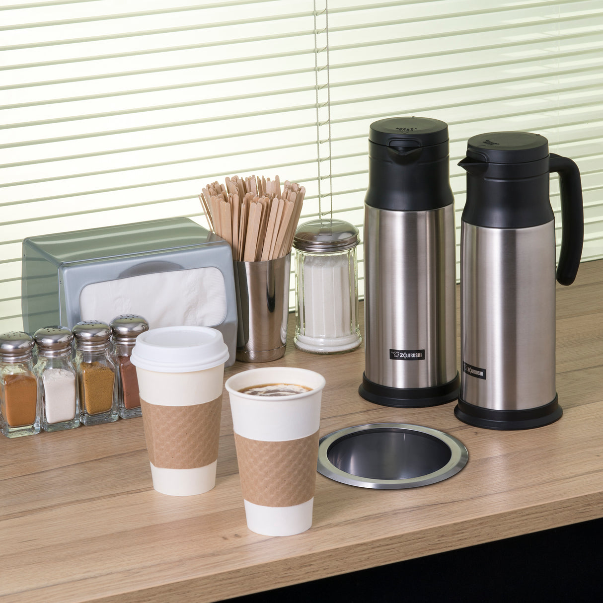A coffee station on a wooden counter features two disposable cups filled with coffee, two stainless steel beverage servers with vacuum insulation, napkins, stir sticks, a sugar jar, and five spice shakers. Horizontal blinds cover the window in the background.