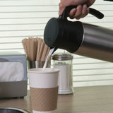 A hand pours milk from a stainless steel beverage server into a paper cup with a brown sleeve. On the counter are wooden stir sticks, a sugar dispenser, a napkin holder, and a window with blinds in the background.