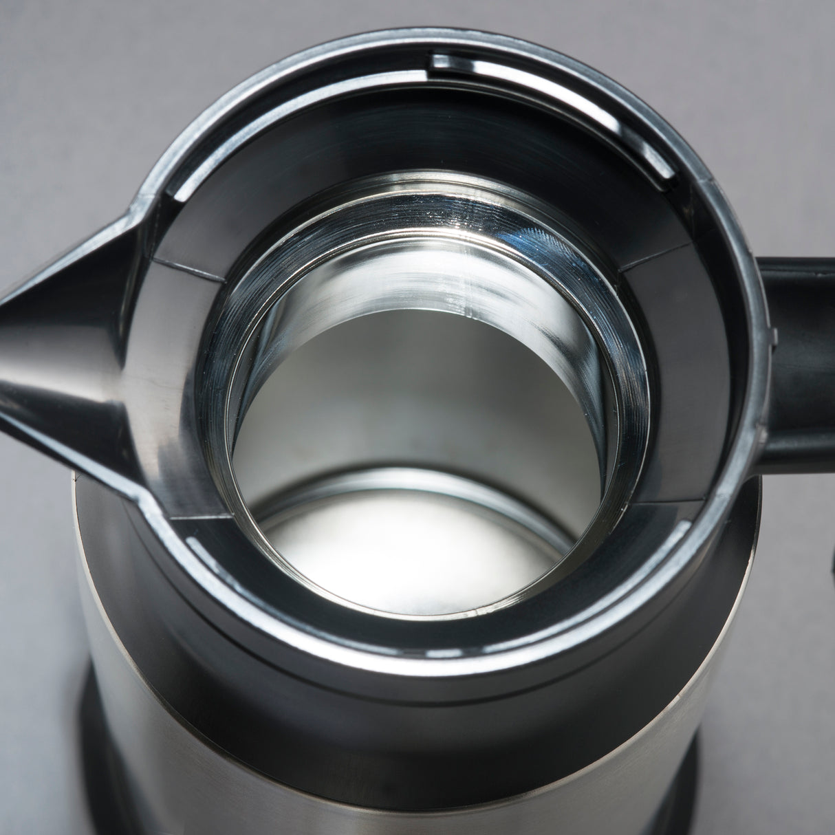 Close-up view looking straight down into the open top of a stainless steel beverage server. The interior is clean and reflective, with a spout on the left and a handle partially visible on the right. Background is plain gray.