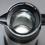 Close-up view looking straight down into the open top of a stainless steel beverage server. The interior is clean and reflective, with a spout on the left and a handle partially visible on the right. Background is plain gray.