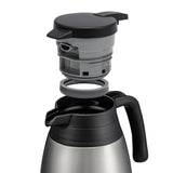 A stainless steel beverage server with a black handle and open mouth. Above the carafe, a gray and black lid and gray sealing ring are separated, illustrating assembly or cleaning. Designed for optimal heat retention. Background is white.