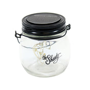A clear glass lantern jar by TableCraft Products with the Shanty logo, featuring a string of LED lights inside. The black lid includes a small solar panel and metal clasps. This LED Solar Lantern sits against a plain white background.