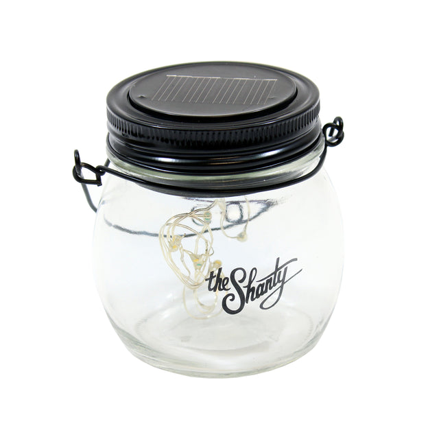 A clear glass lantern jar by TableCraft Products with the Shanty logo, featuring a string of LED lights inside. The black lid includes a small solar panel and metal clasps. This LED Solar Lantern sits against a plain white background.