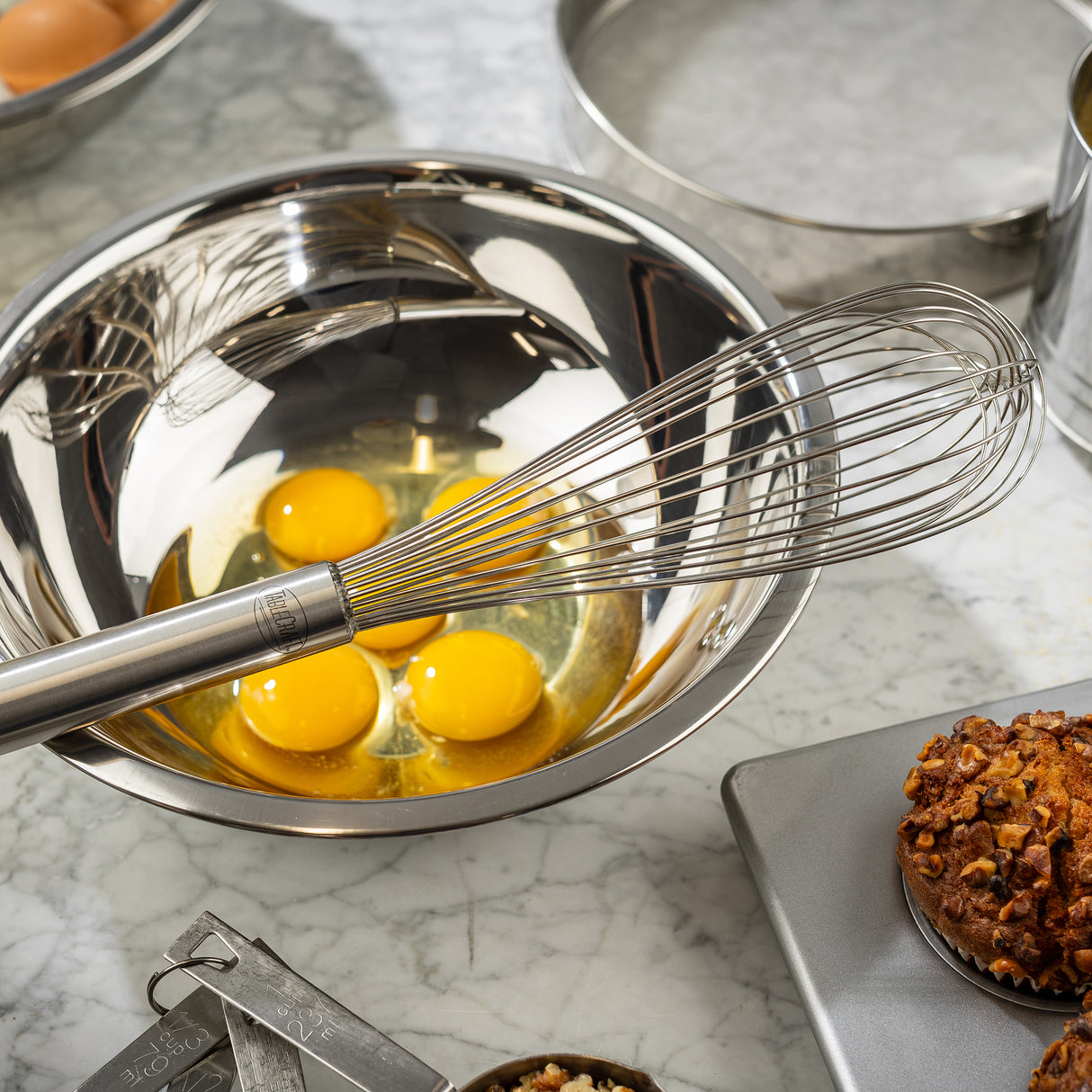 A TableCraft Piano Whip rests in a shiny stainless steel bowl containing three cracked eggs on a marble countertop. Nearby are measuring spoons, part of a muffin on a scale, and other baking tools reflecting the bright kitchen lighting.