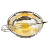 A TableCraft Piano Whip rests in a shiny stainless steel mixing bowl containing a mound of yellow cornmeal with a single cracked egg in the center, surrounded by white sugar, set against a white background.