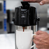 A hand presses the top of a black and silver Zojirushi Airpot with a stainless steel vacuum liner, dispensing coffee into a white mug in a kitchen or breakroom, ensuring optimal heat retention.