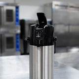 A stainless steel airpot with a black pump lid and stainless steel vacuum liner sits on a metal counter in an industrial kitchen, with stainless steel appliances and ovens blurred in the background for optimal heat retention.