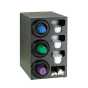 A black cup dispenser cabinet holds three rows of plastic coffee pods in green, blue, and purple, with spaces for white disposable cups and lids on the right. The bottom right compartments contain white coffee stirrers. Stands upright on a white surface.